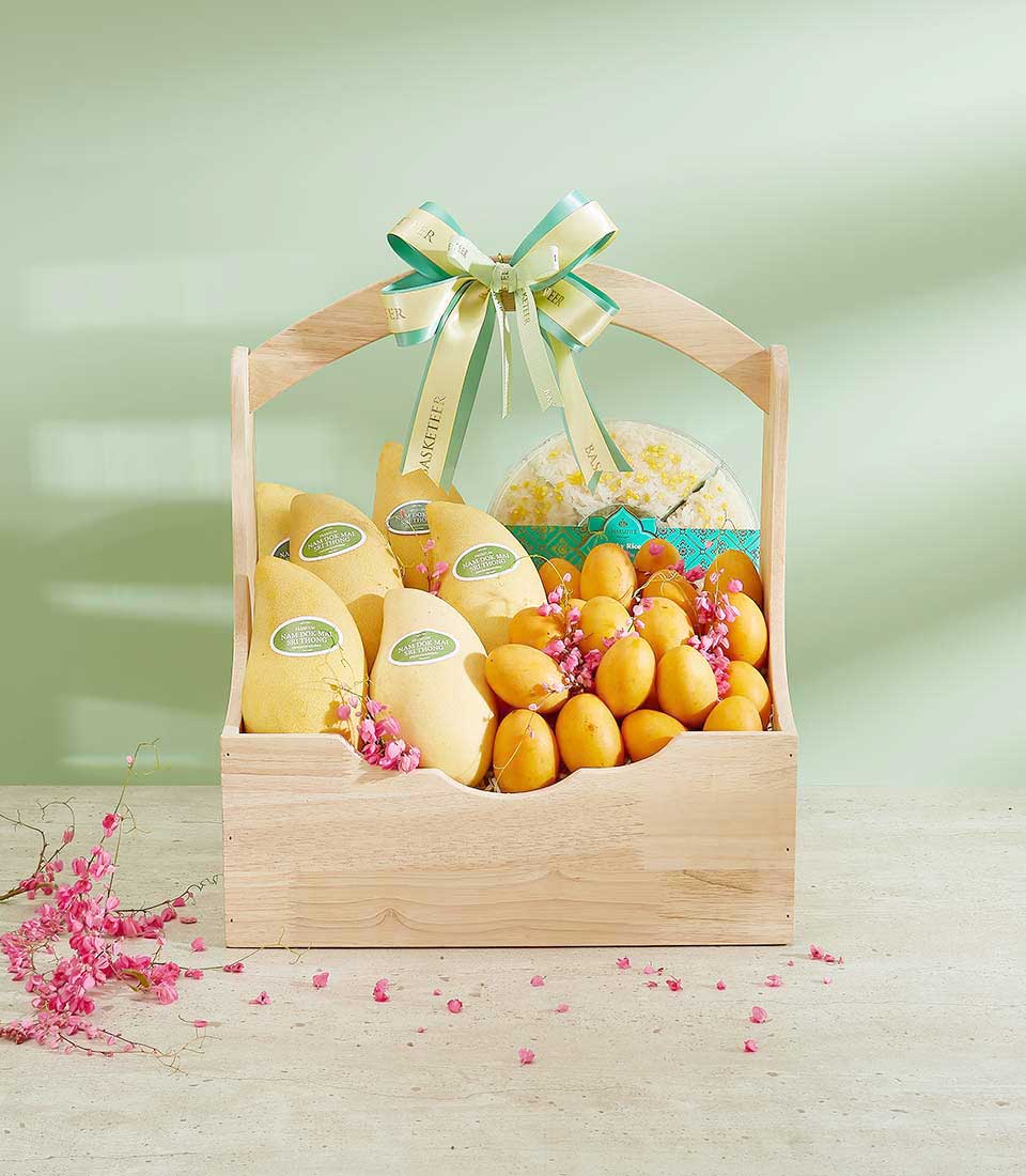 The Yellow Bliss Fruits Basket features a wooden basket filled with yellow mangoes and a mix of other tropical fruits. Adorned with a green and yellow bow, this basket is surrounded by scattered pink flowers at the bottom, set against a light green wall backdrop.