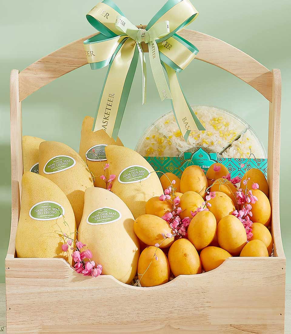 The Yellow Bliss Fruits Basket is a wooden basket filled with ripe yellow mangoes and mini mangoes, decorated with pink flowers. It features a ribbon with text and a circular container of dried fruit. The background is light green, and the basket has a sturdy handle.