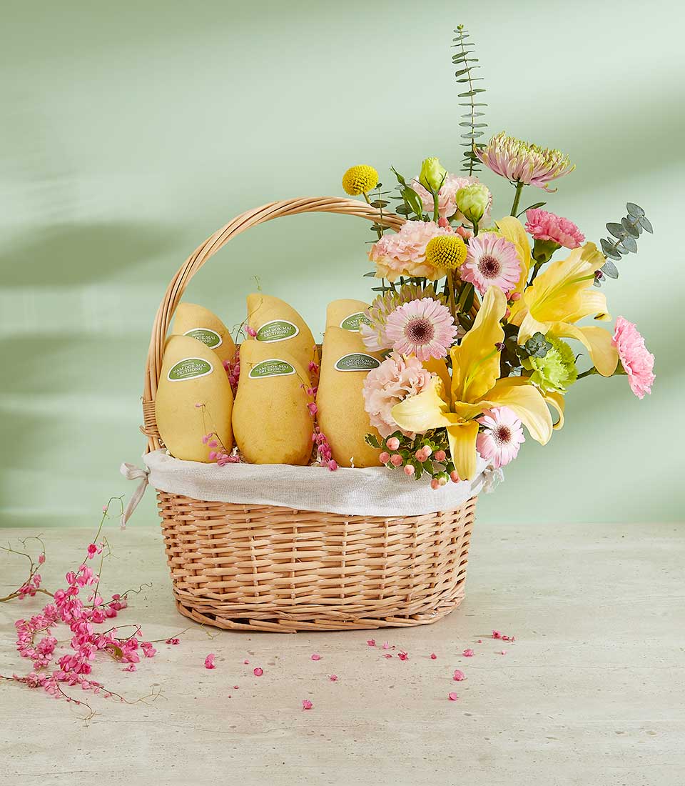 An Authentic Mango Delight Basket features a wicker basket filled with five yellow mangoes on the left side and a vibrant floral arrangement on the right. The arrangement includes yellow lilies, pink carnations, pink gerbera daisies, eucalyptus, and small yellow spherical flowers, set against a light green background.