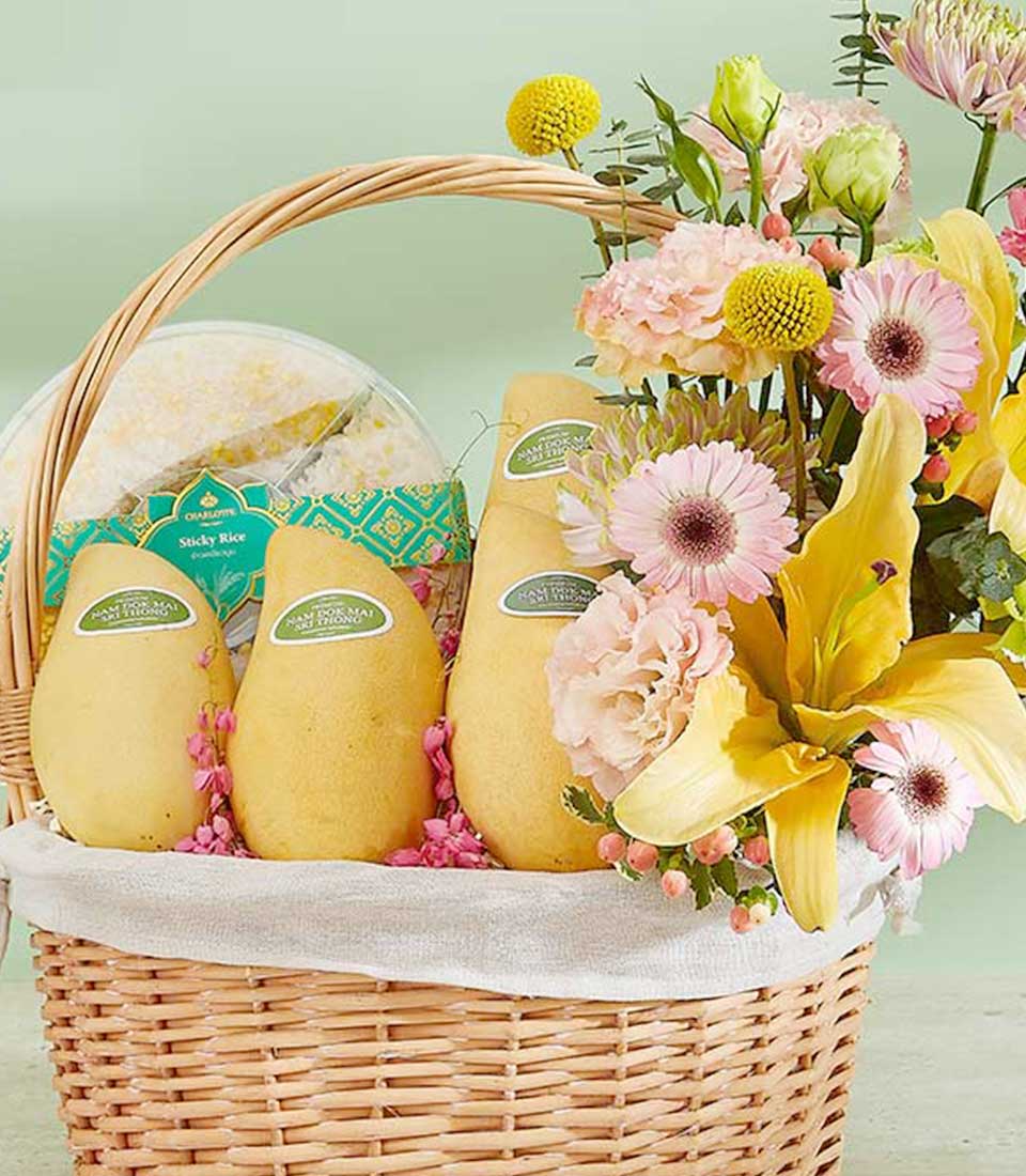 The Timeless Mango Basket features a wicker basket filled with yellow mangoes, pink and yellow flowers, and a package of sticky rice. Displayed against a light green background, the arrangement is beautifully lined with a white cloth.