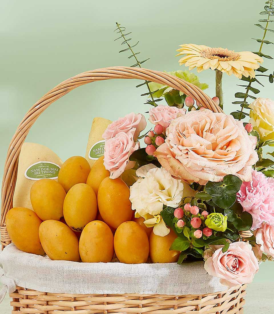 An opulent Thai fruit elegance is showcased in a wicker basket filled with yellow mangoes and an assortment of pink, white, and yellow flowers, including roses and gerberas, arranged against a soft green background. The basket has a white cloth lining and is adorned with greenery.