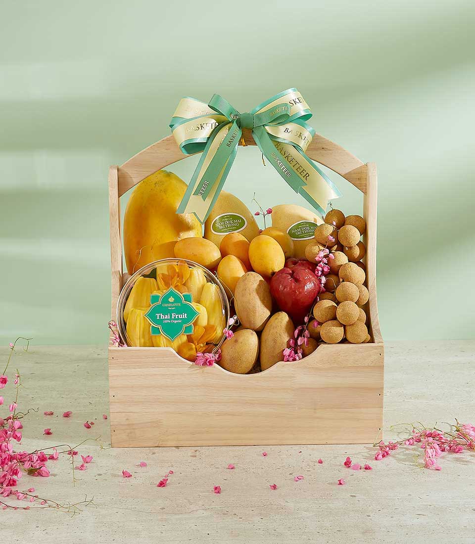 An Elegant Tropical Fusion Gift Basket featuring assorted fruits, including a whole yellow mango, lychees, longans, and another red fruit. Adorned with green and white ribbon bows, the wooden basket is beautifully decorated with pink flowers scattered around its base.