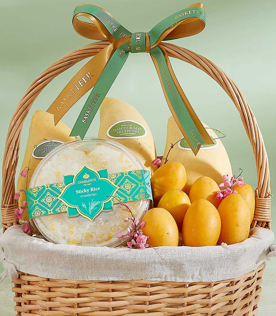 The Thai Harvest Delight Basket features a wicker basket filled with yellow mangoes, two packets of crispy coconut rolls, and a container of sticky rice. Adorned with a green and yellow ribbon, it sits on a light-colored cloth against a soft green background.