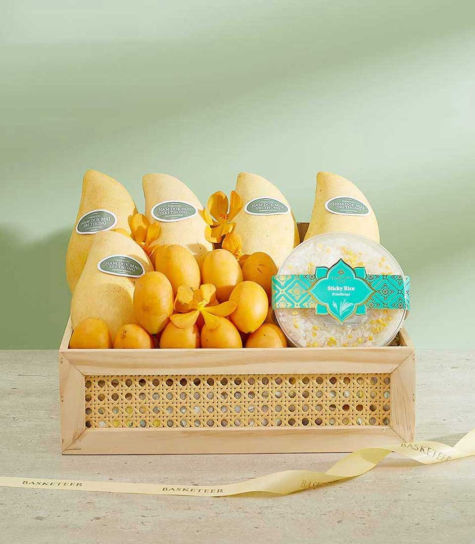 A wooden tray filled with neatly arranged yellow mangoes with labeled stickers, small yellow fruits among yellow flower decorations, and a hexagonal green package of sticky rice. The exquisite Summer Fruits Delight is set against a light green background with a yellow ribbon draped nearby.