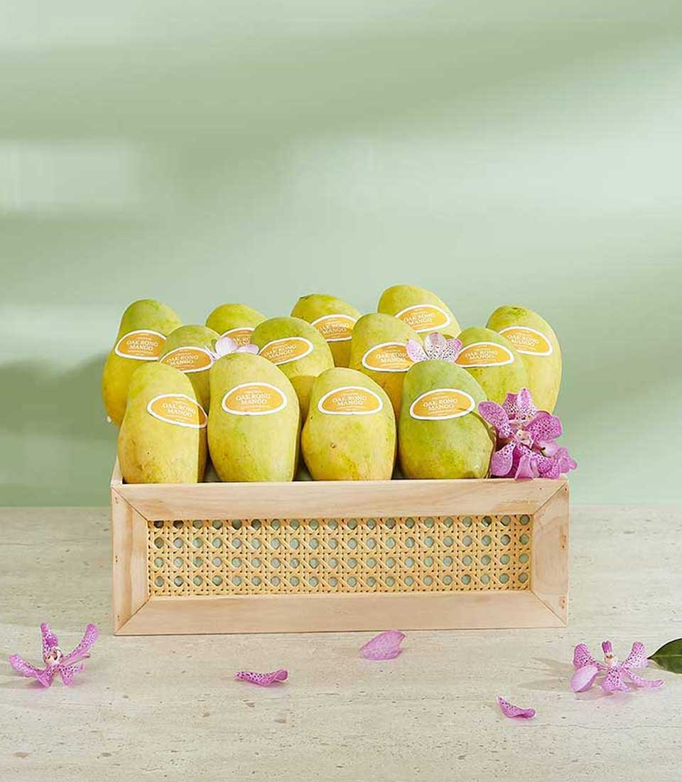 A wooden crate filled with ripe Yellow Mangoes, each bearing a golden label, is placed on a light wood surface. There are pink orchid flowers scattered around the crate. This Oak Rong Mango Gift Box sits against a soft green wall, making it a delightful and elegant presentation.