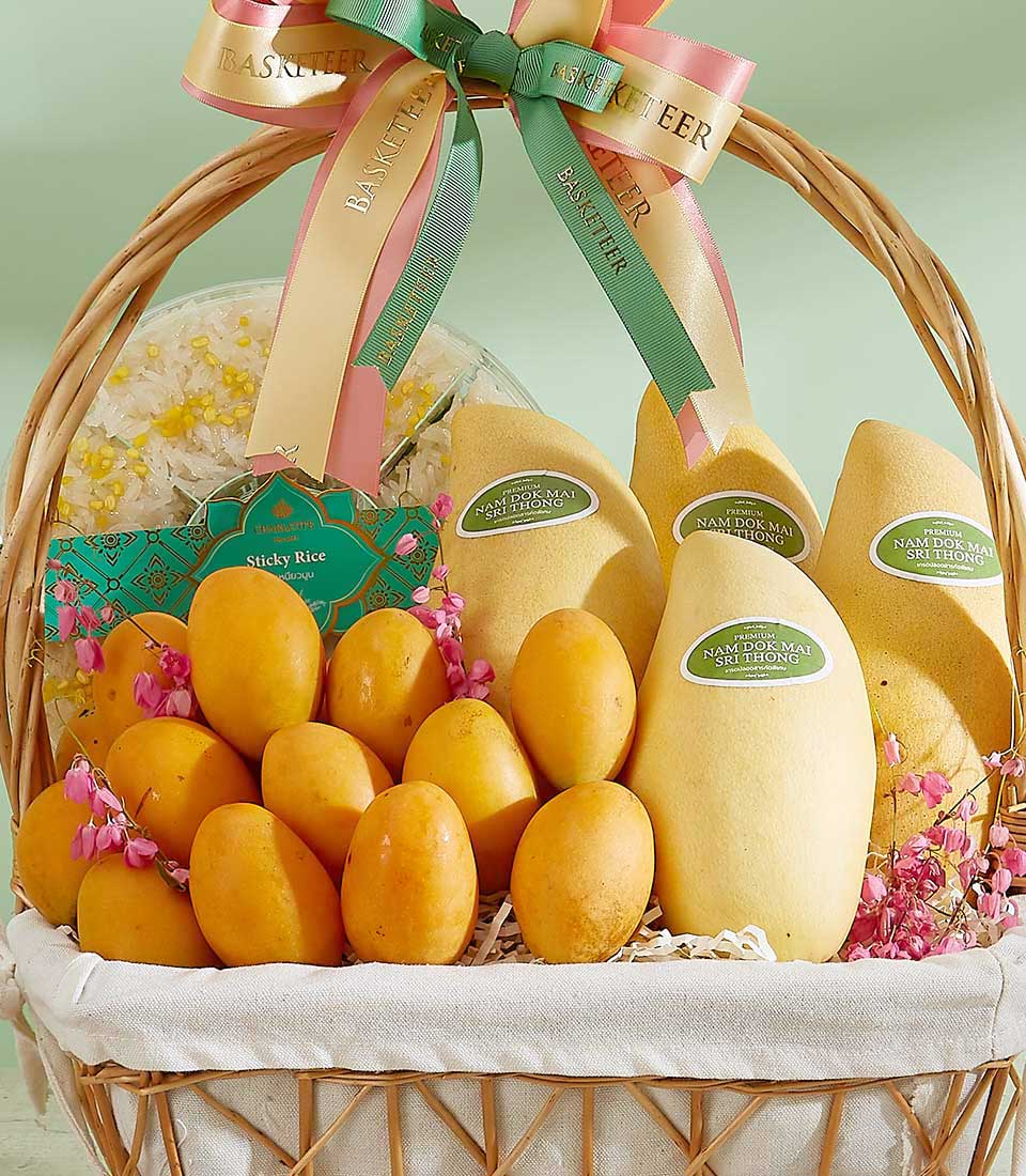 The Tropical Fruits Harmony Basket features a wicker basket filled with golden mangoes and small orange fruits, adorned with pink flowers and ribbons. It includes a package of sticky rice and green-labeled mangoes. The ribbons have 