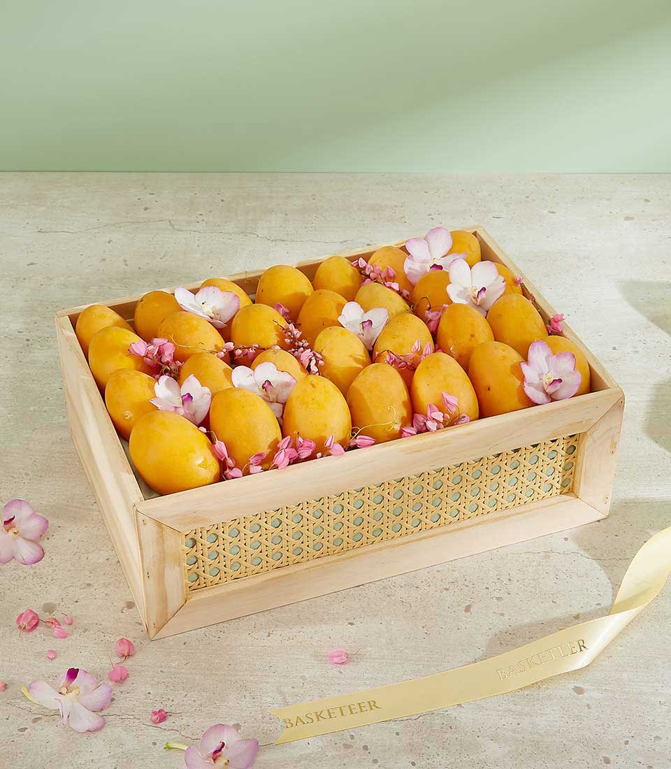A wooden tray filled with ripe, golden mangoes, decorated with pink flower petals and scattered orchid blossoms. The tray has a woven design on the sides and is placed on a light-colored surface. A ribbon with 