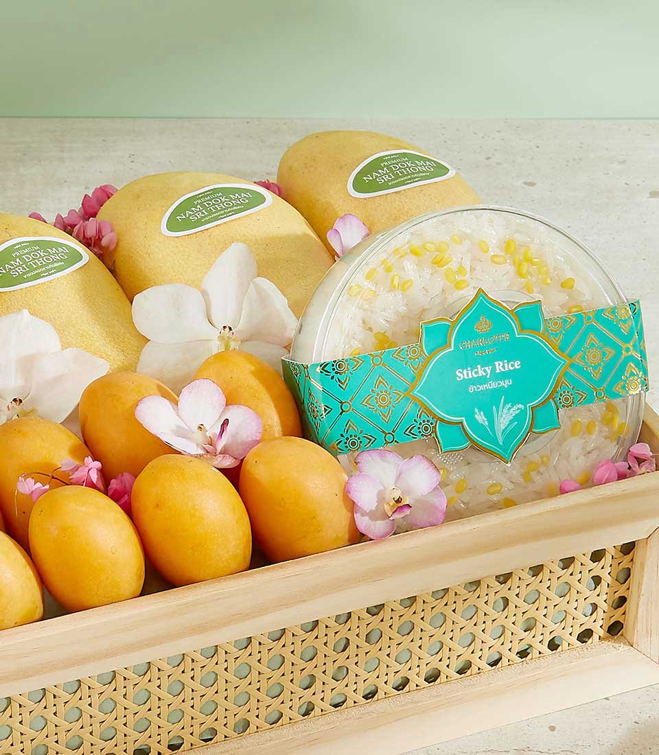 A wicker tray containing three whole ripe mangoes, several smaller yellow fruits, and a bowl of sticky rice, garnished with floral decorations. The teal and gold label on the sticky rice container reads 