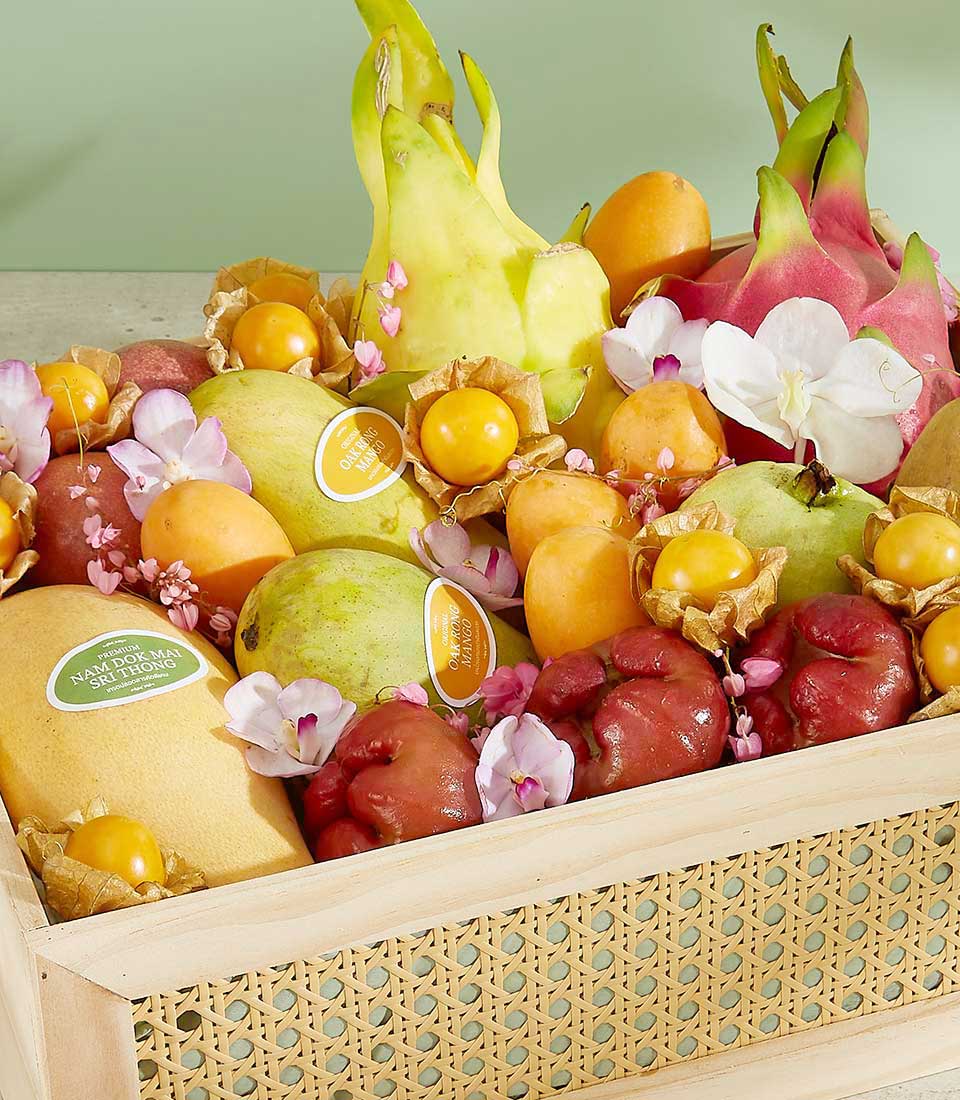 A wooden basket filled with a colorful variety of tropical fruits, including dragon fruits, mangoes, papayas, cape gooseberries, and others. This Exquisite Thai Summer Fruits Box is decorated with delicate white and pink orchid flowers.