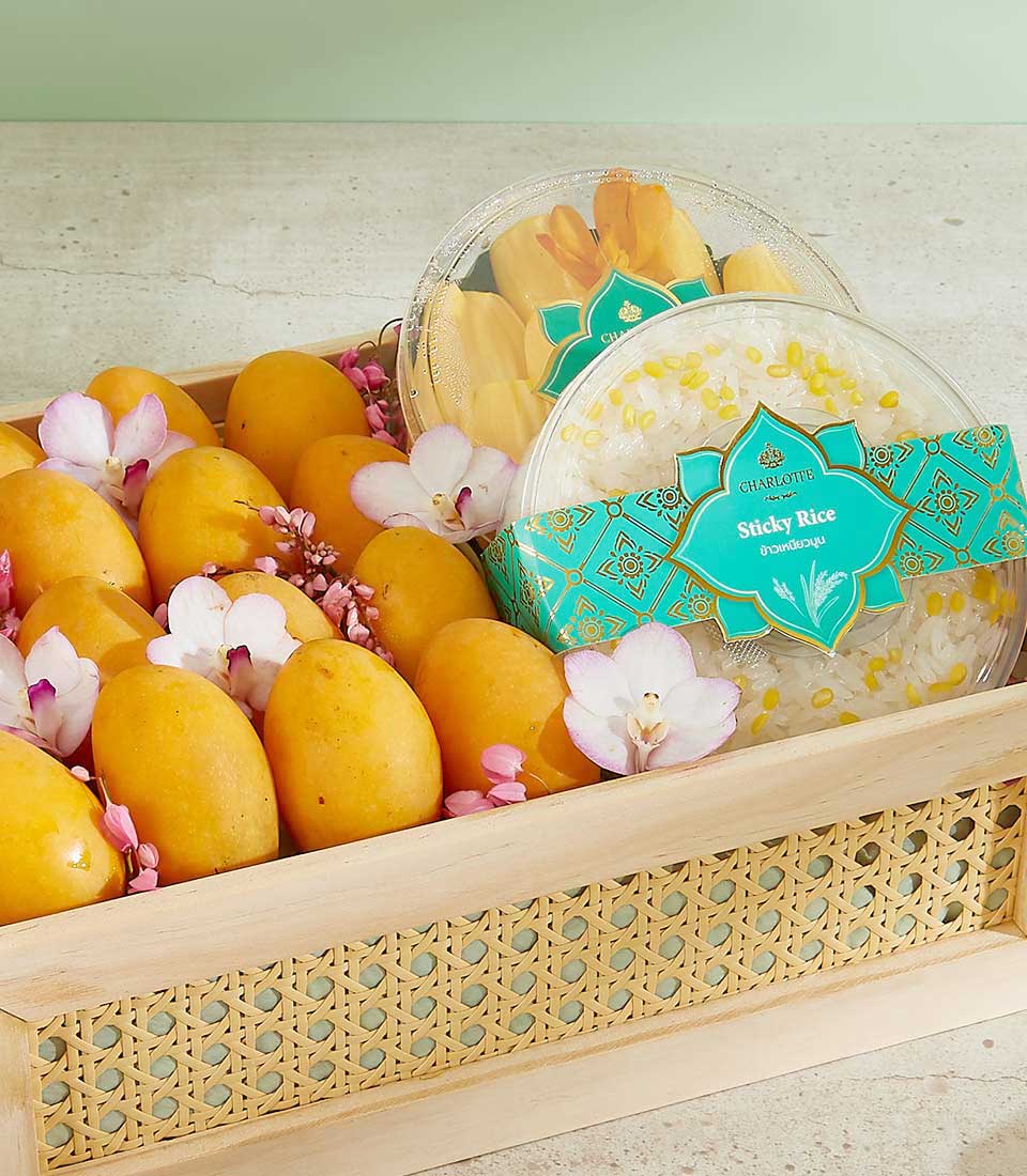 An Authentic Mayongchid Gift Crate holds a wooden basket filled with ripe yellow mangoes, pink and white flowers, and two plastic containers of mango sticky rice wrapped with turquoise labels. The sticky rice containers are decorated with patterns and labeled 