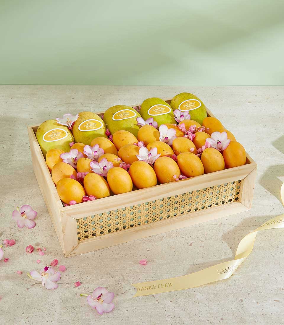 A rectangular wooden basket containing neatly arranged mangoes, some with greenish-yellow skin and others more orange, adorned with pink artificial flowers, sits on a beige surface. Beside the basket lies a cream-colored ribbon with 