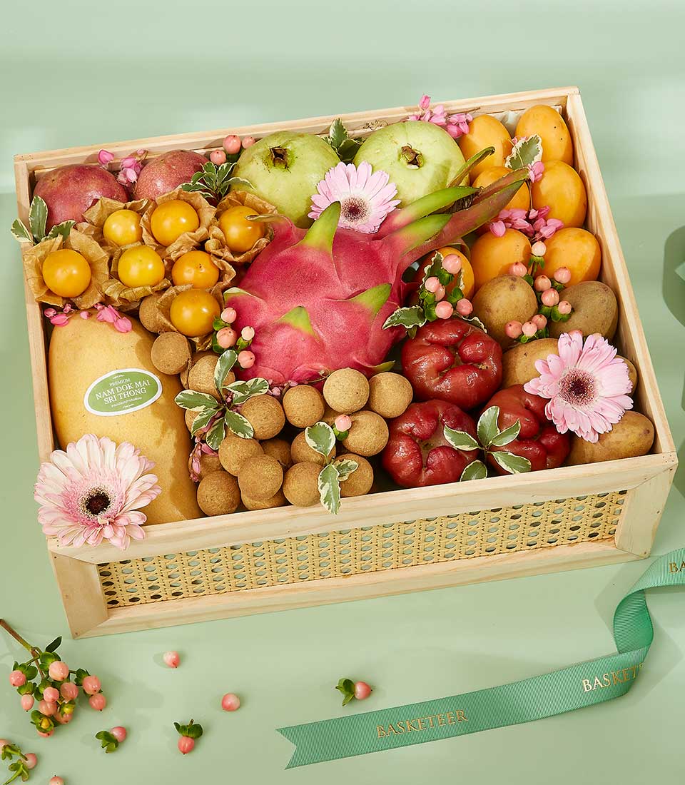 A wooden crate filled with an assortment of fruits including Thai fruits, apples, dragon fruit, kiwis, and berries, adorned with pink flowers and green leaves. The crate is set against a pastel green background with a green ribbon labeled 