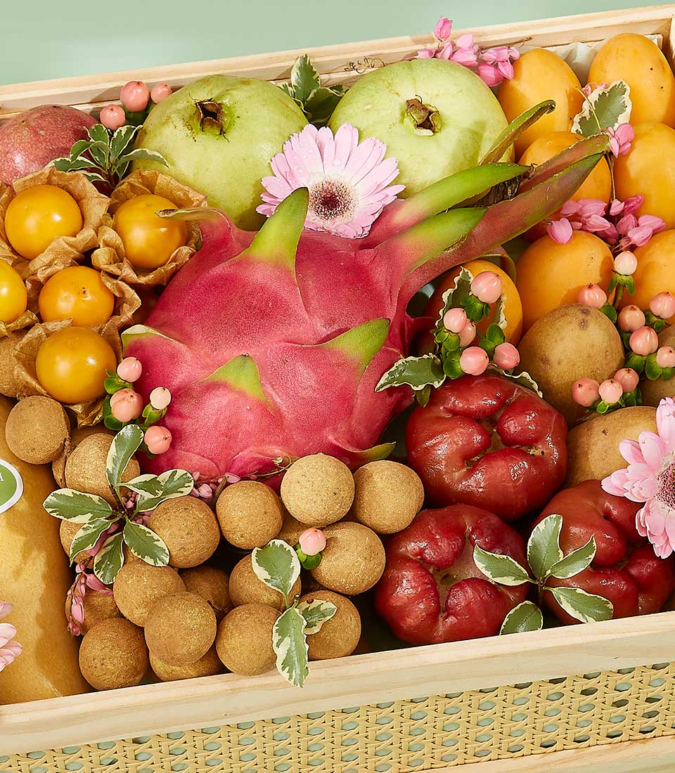 A wooden crate filled with a colorful assortment of Thai fruits, including a prominent dragon fruit at the center, surrounded by green apples, lychees, yellow gooseberries, bananas, and red rose apples. The arrangement is accented with pink flowers and green leaves.