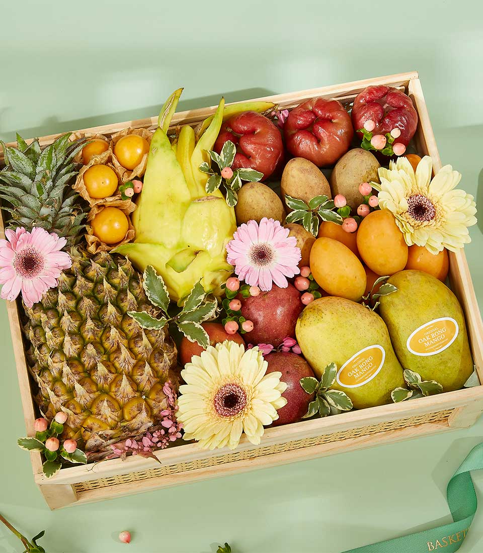 A wooden box sits on a mint-green surface filled with a variety of vibrant fruits, including pineapple, papaya, mangoes, apples, and kiwifruit. Interspersed are pink and yellow gerbera daisies adding a decorative touch. This elegant tropical Thai gift is finished with a green ribbon partially visible on the right.