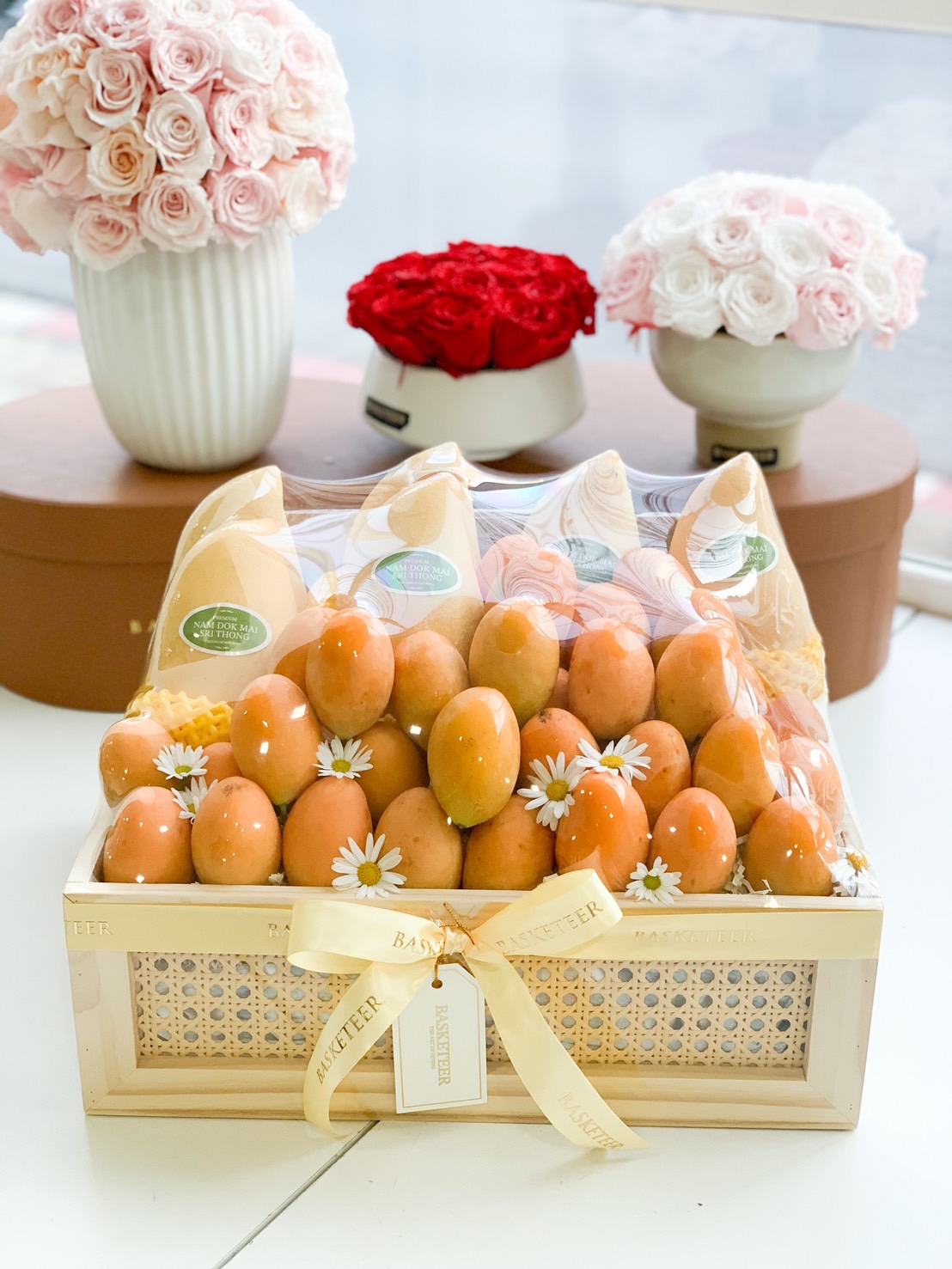 A luxurious fruit gift basket featuring premium Mayongchid (Marian Plum) and Nam Dok Mai See Thong mangoes, elegantly arranged in a handcrafted wooden crate with delicate floral decorations. Wrapped with a golden ribbon and a personalized Basketeer tag, this exquisite fruit box makes for a perfect luxury gift for any occasion.