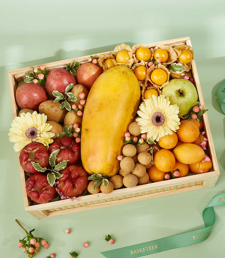 An enchanting mixed fruit basket, featuring a wicker tray filled with assorted fruits including a large papaya, green apple, red apples, yellow and orange delights, and small brown berries. The tray is beautifully decorated with yellow and red flowers, along with small pink berries on a light green surface.