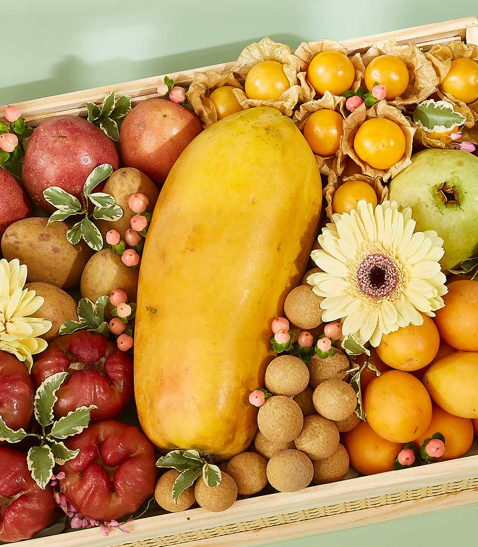 An enchanting mixed fruit basket filled with assorted fruits, including a large yellow mango, red apples, green apples, longans, and golden berries, adorned with pink and white flowers and green leaves. The arrangement is colorful and visually appealing.