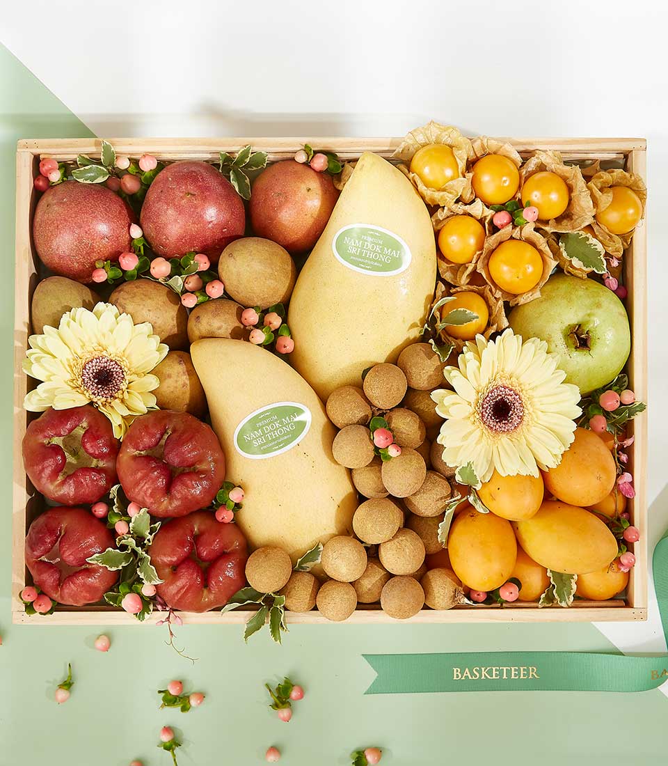 A wooden tray filled with an assortment of fruits including apples, mangoes, lychees, longans, and yellow cherry tomatoes is beautifully decorated with two yellow gerbera daisies and green foliage. The tray features a 