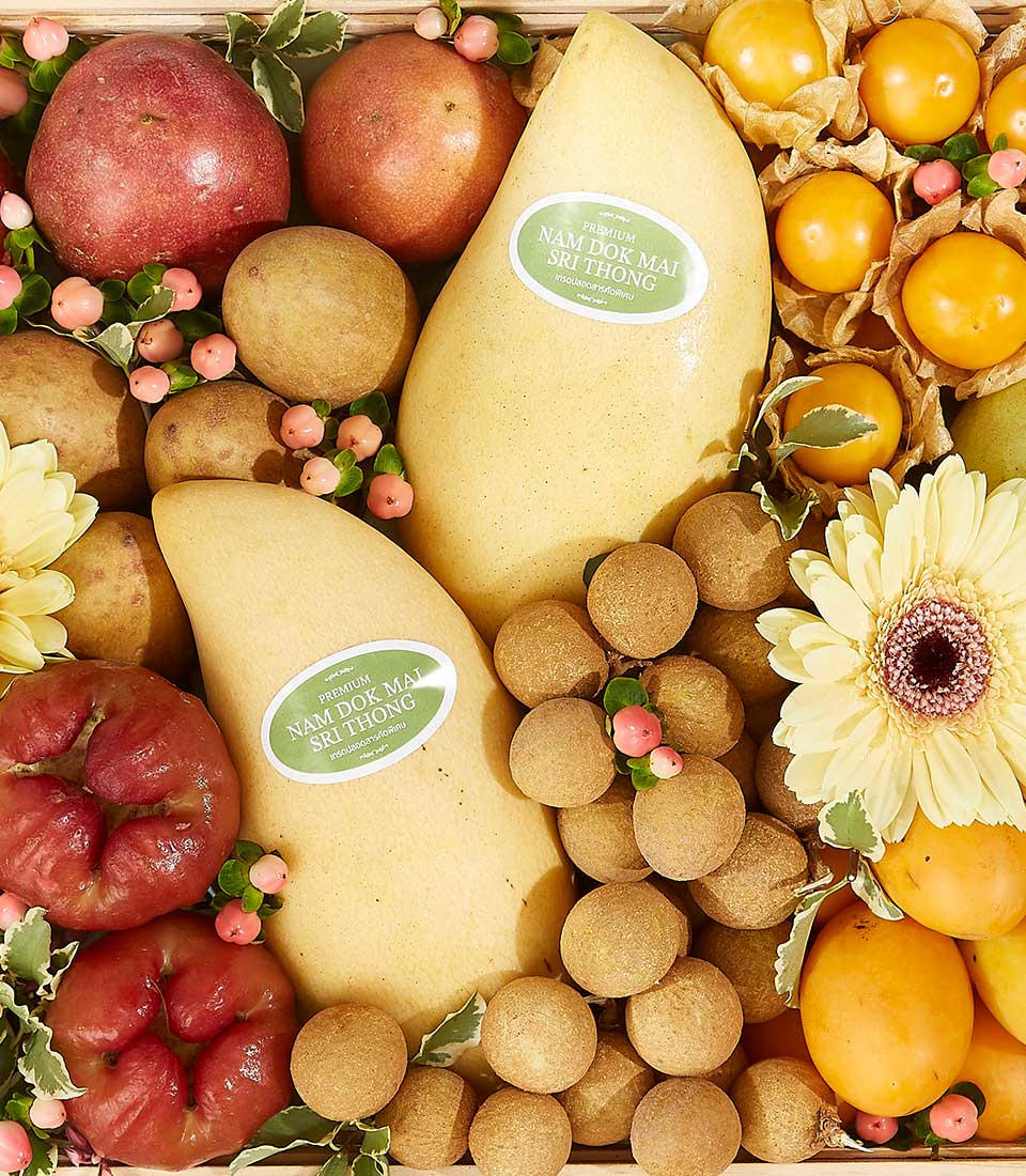 A Tropical Harvest Fruit Basket in a wooden crate, featuring two large yellow mangoes with labels, surrounded by red apples, orange persimmons, kumquats, lychees, yellow daisies, and small decorative berries on green leaves.