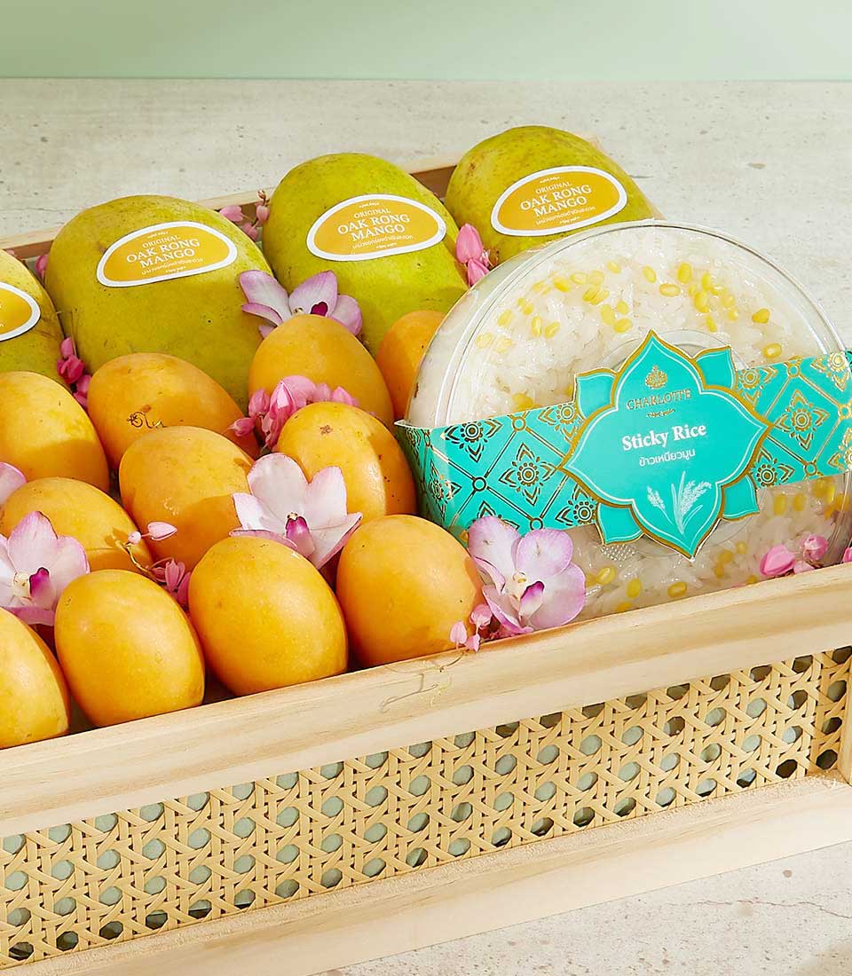 A wooden tray holds several splendid green and yellow mangoes with labels, surrounded by pink and white blossoms. Next to the mangoes, there is a package labeled 