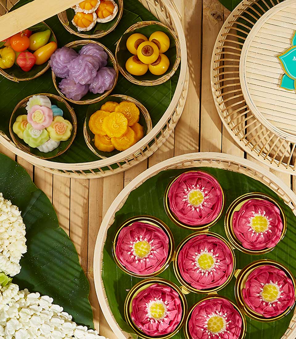 An overhead view of a vibrant Thai sweet dessert assortment arranged in bamboo baskets. The display includes intricately shaped red floral desserts, colorful fruit-shaped sweets, and purple, green, and yellow treats. White jasmine flowers lie on the left side of the bamboo mat.