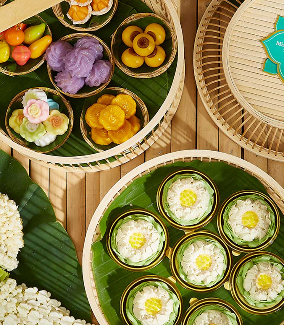 An assortment of beautifully crafted Thai desserts displayed in bamboo baskets on a green banana leaf. This Thai Dessert Medley Basket features colorful treats including floral-shaped sweets, fruit-shaped candies, and intricate molds, showcasing a vibrant and appetizing presentation.