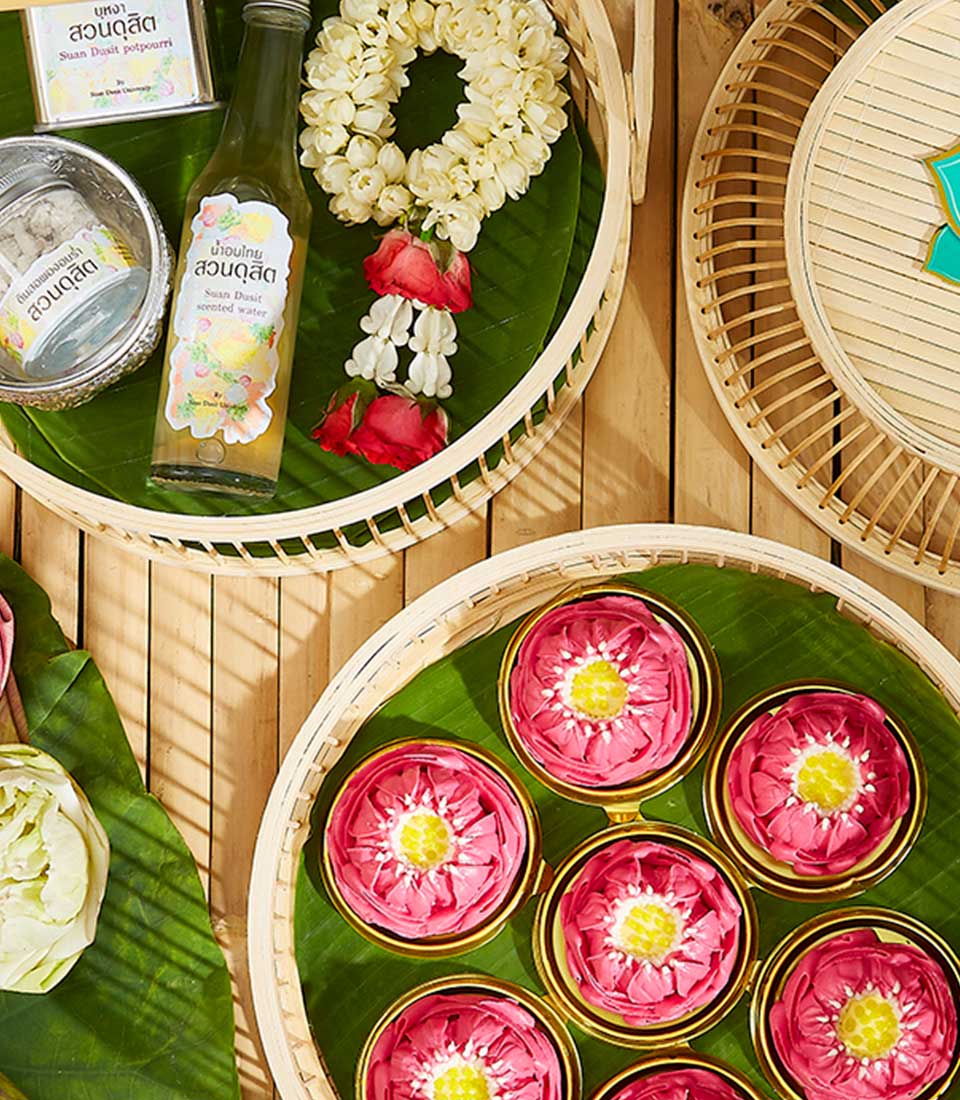 A top-view of the Thai New Year Delight Set arranged on a bamboo mat. There are baskets containing pink lotus flowers, a bottle of Thai beverage, a garland of white flowers with red petals, and a container of sweets. The items are displayed on green banana leaves.