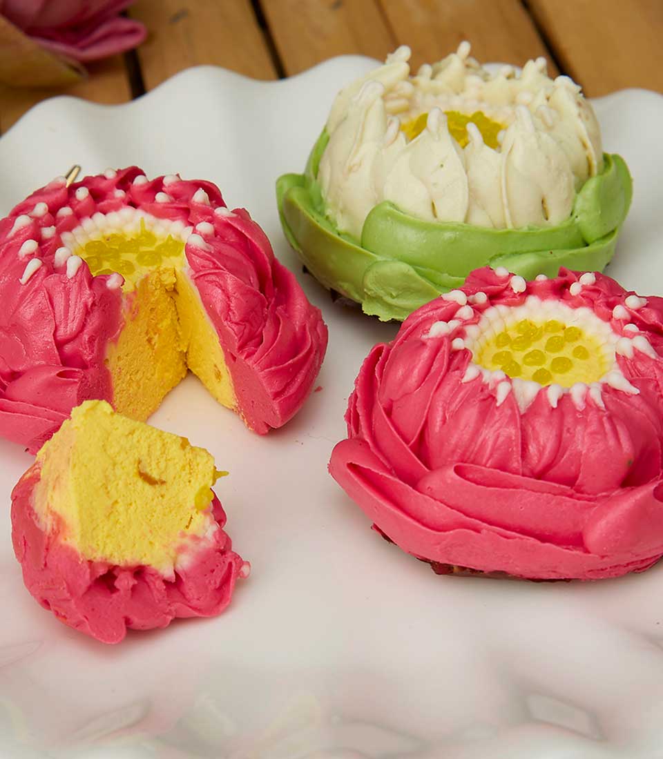 A plate showcases three flower-shaped desserts, each resembling a lotus. Two are pink with yellow centers and one is white with a green outer layer. One pink dessert is cut open, revealing a yellow filling inside. These Summertime Lotus Treats rest beautifully on a white, scalloped-edged plate.