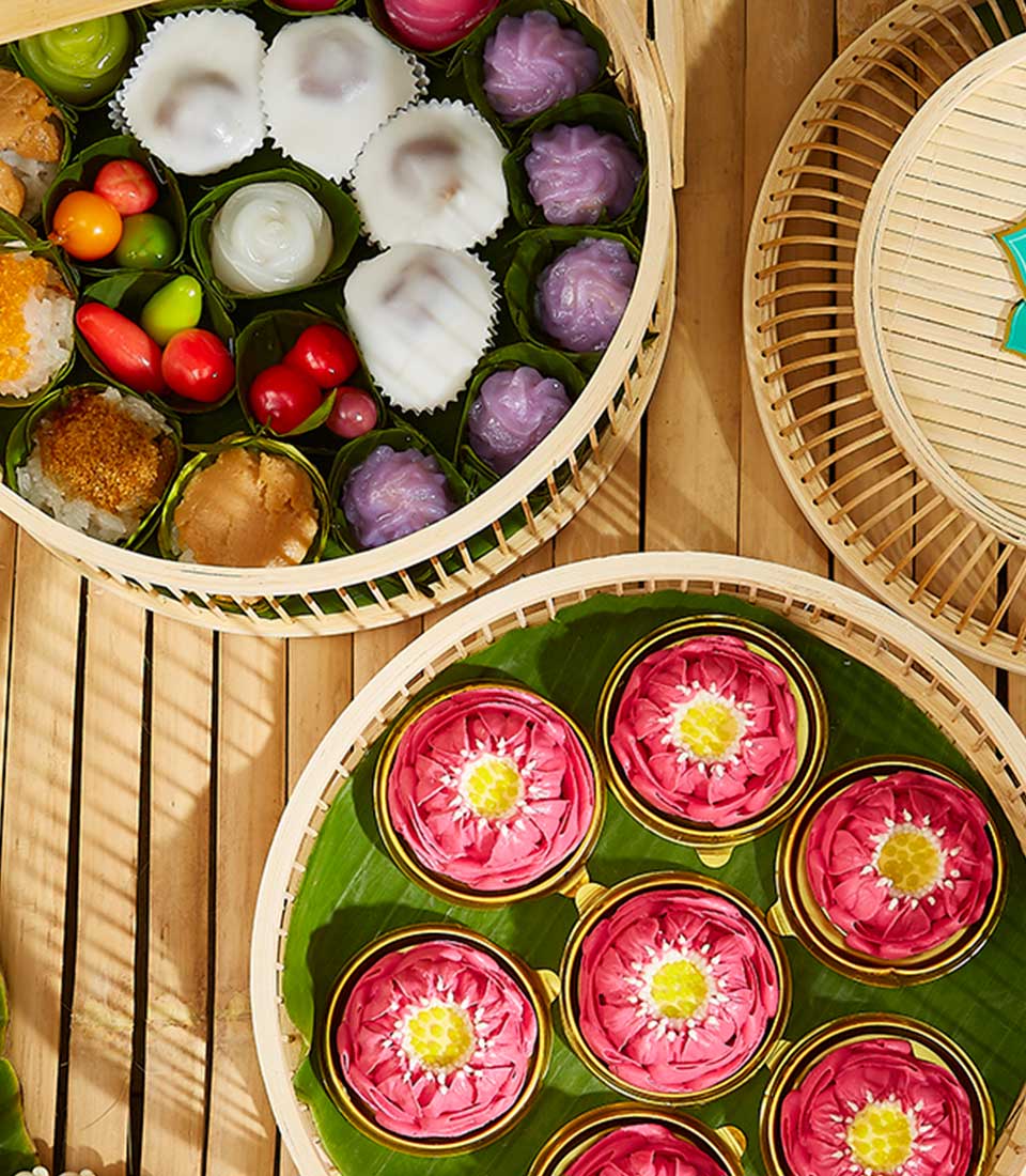 Top view of a colorful assortment of dim sum in round bamboo steamers. One steamer holds an array of dumplings and small vegetables, while another is filled with pink flower-shaped dim sum reminiscent of a Thai Songkran Dessert Basket. The third shows an open steamer lid on a bamboo mat background.