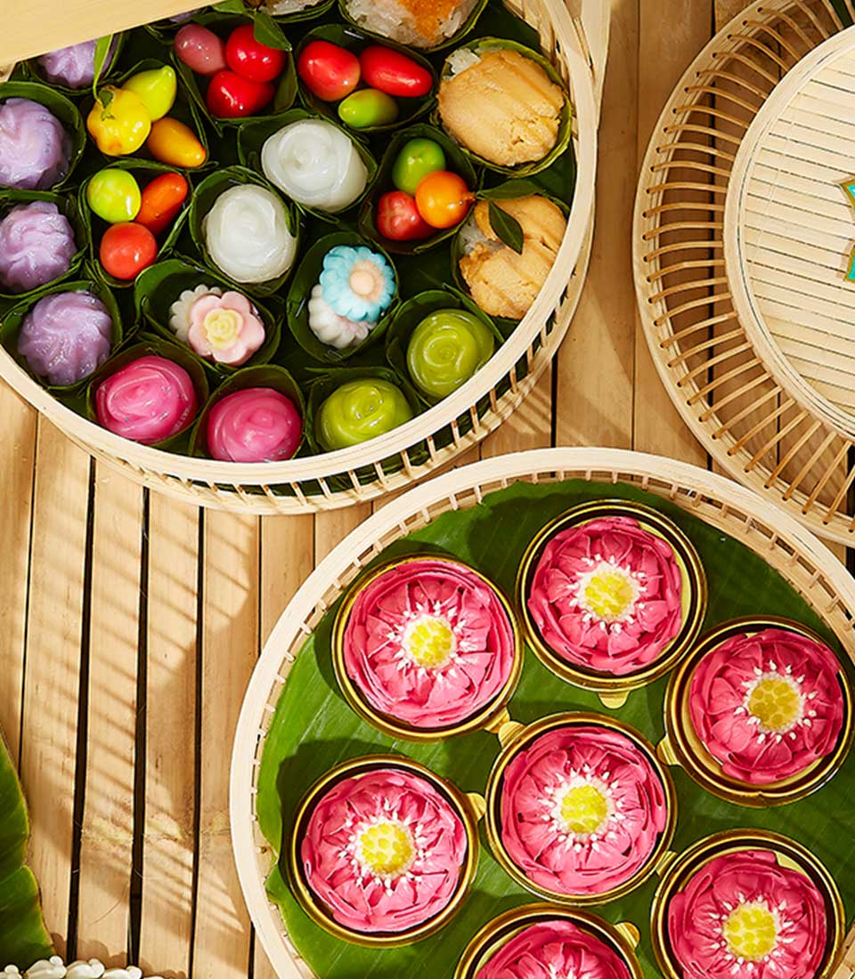 A variety of colorful, artistically crafted Thai desserts neatly arranged in Luxe Thai Dessert Baskets. The desserts include fruit-shaped sweets, flower-shaped jellies, and intricately molded treats, all displayed on green leaves against a wooden backdrop.