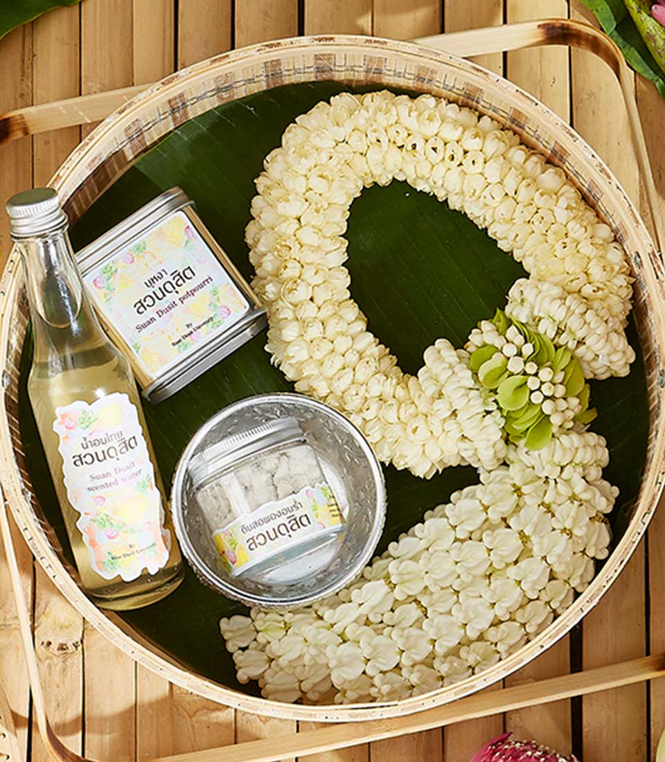 A woven tray holds a fragrant Songkran garland of white flowers, a clear glass bottle with a floral label, and a small tin alongside a smaller plastic container. All items rest on a leaf atop the bamboo surface.