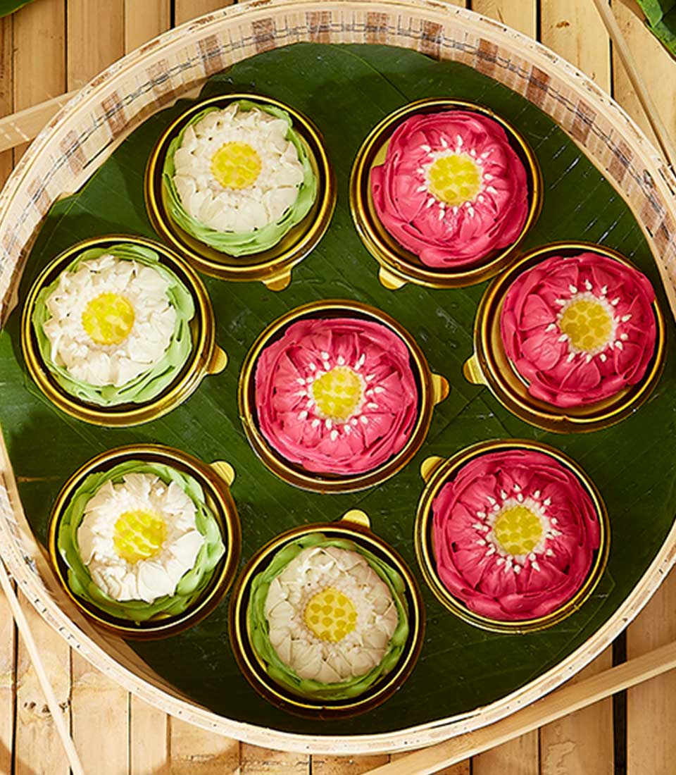 A round bamboo basket lined with green leaves contains eight intricately folded lotus flower arrangements. The flowers, resembling pink and white lotus jelly candies with yellow centers, are placed in neat rows within golden bases. This unique gift rests on a wooden surface.