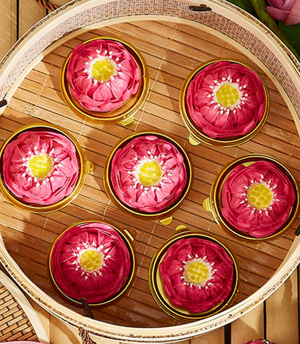 Top view of a round bamboo steamer basket containing six pink lotus-shaped Thai jelly candies with yellow centers, arranged symmetrically. The intricate design and vibrant colors of these artisanal treats mimic the appearance of real lotus flowers, making it a perfect gift.