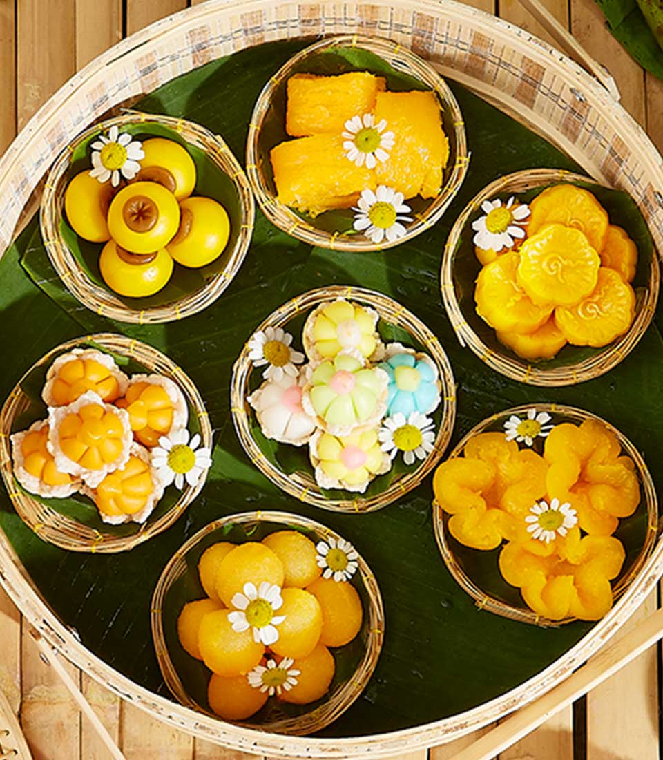 A delightful Thai Dessert Surprise Gift: a basket containing seven small woven trays of colorful traditional Thai desserts, beautifully arranged on a green banana leaf. The desserts are adorned with small white and yellow flower decorations, adding a festive touch to the presentation.