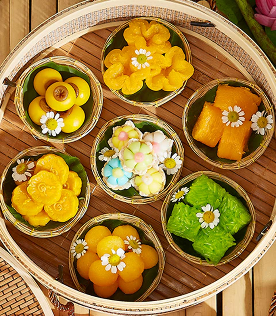 A round woven tray holds seven smaller woven baskets, each filled with a variety of colorful Thai desserts. The Sweet Bliss Dessert Gift is arranged attractively and garnished with small white flowers. The wooden table underneath adds a rustic touch.