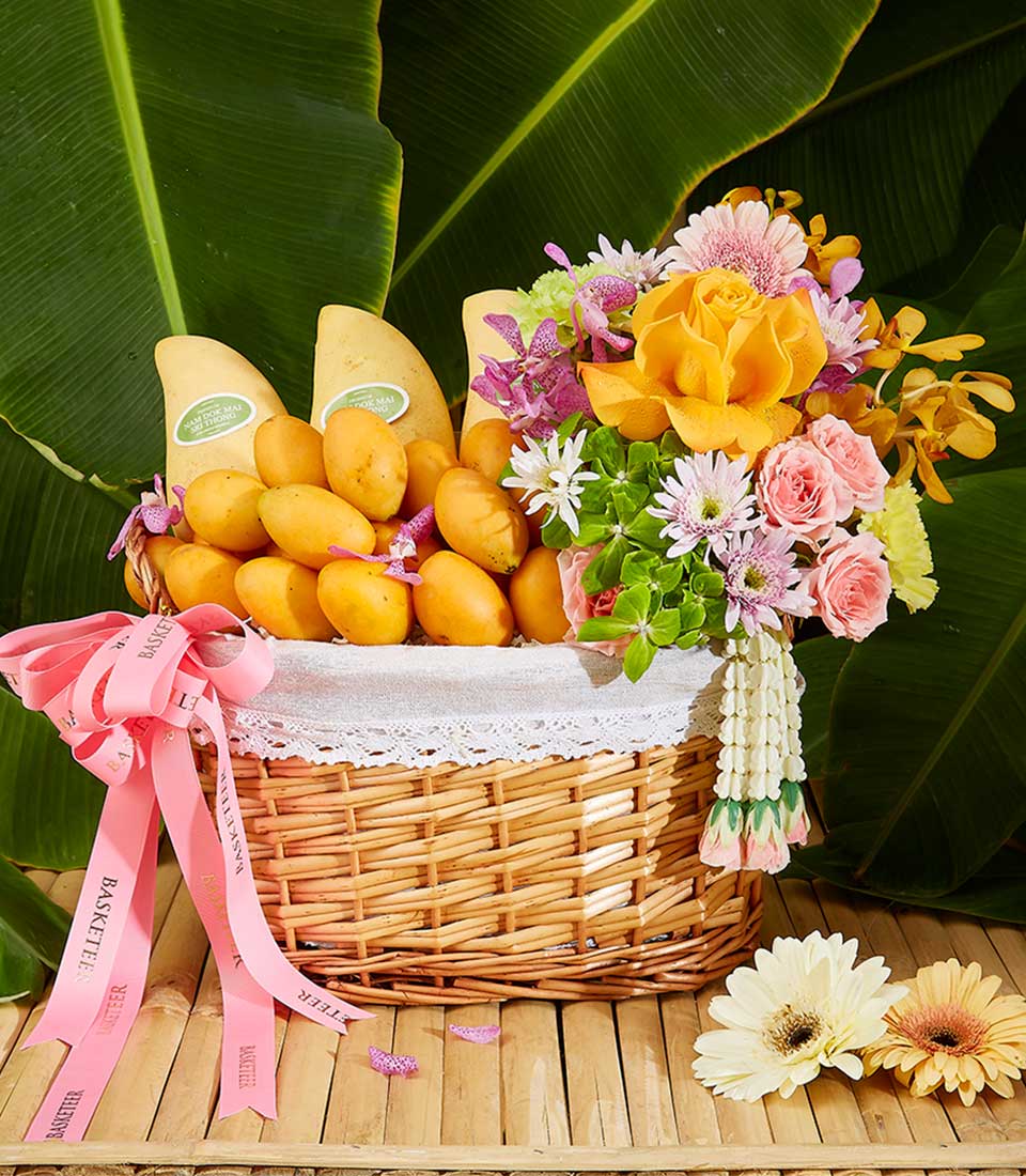 A woven basket filled with ripe yellow mangoes and a colorful assortment of flowers, including roses and daisies. The Tropical Songkran Delight Basket is adorned with pink ribbons and set against a backdrop of large green leaves, resting on a wooden surface with additional flowers nearby.