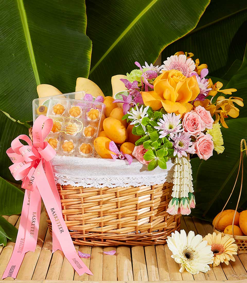 A wicker basket filled with yellow mangoes, colorful flowers including roses and chrysanthemums, and a box of confections. The Tropical Songkran Celebration Hamper is adorned with pink ribbons and set against a backdrop of large green leaves.