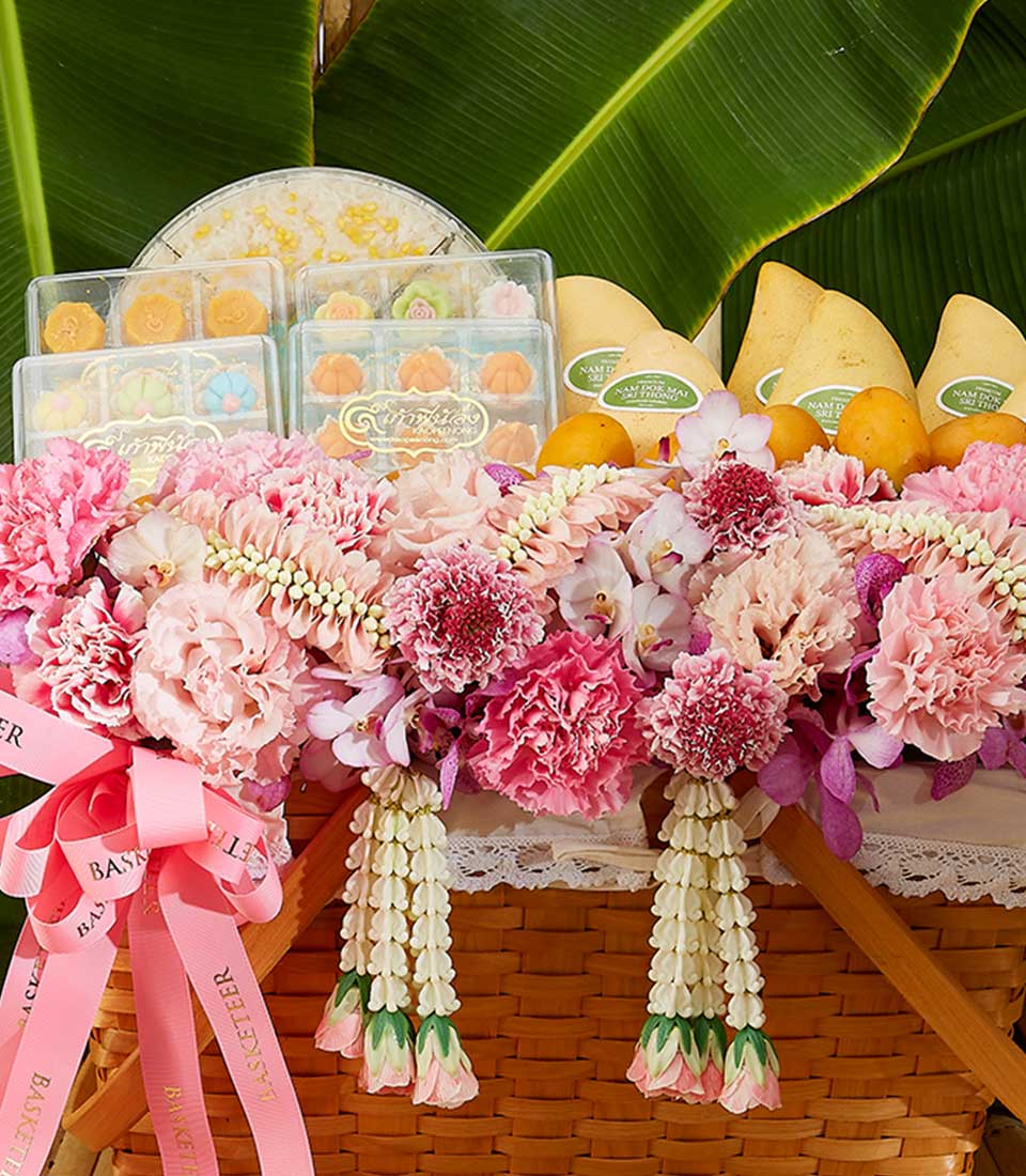 A Thai Blossom Summertime Basket features a wicker container brimming with pink and white flowers, accented by pink ribbons, surrounded by boxes of sweets and vibrant yellow mangoes. The basket's handles are adorned with floral garlands, while large green leaves form the backdrop.