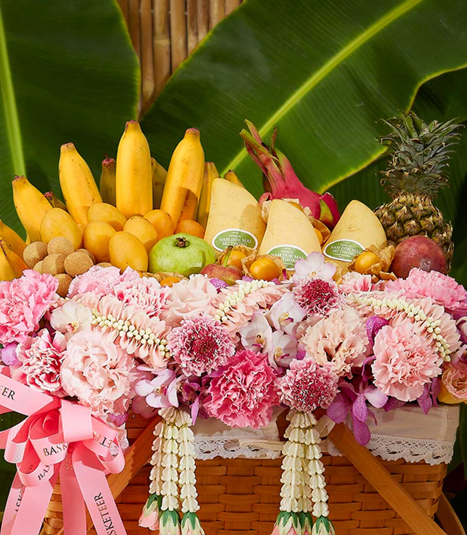 A Tropical Songkran Fruits Basket brimming with fresh tropical fruits including bananas, dragon fruit, pears, pineapple, and more. The wicker basket is adorned with pink carnations, orchids, and decorative ribbons. Lush green leaves serve as a beautiful backdrop.