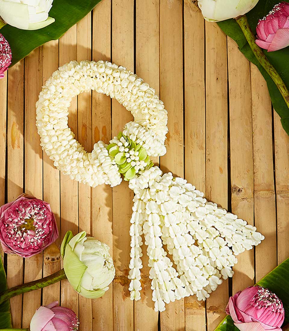 A circular white floral garland gift, adorned with green accents, is laid out on a wooden surface surrounded by flowers in shades of white, pink, and purple. The garland displays a cascading fringe of small white blossoms amidst partially and fully blooming flowers.