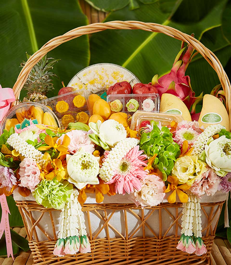 A wicker basket brimming with an assortment of colorful fruits, flowers, and various small packages. Tropical fruits like dragon fruit and pineapples are visible, surrounded by fresh vibrant flowers and greenery. The background features large green leaves, evoking the charm of an elegant Thai festival gift.