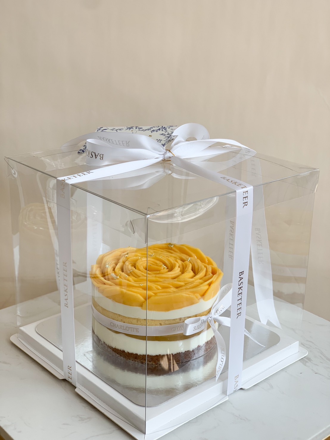 A luxurious mango cheesecake elegantly packaged in a clear gift box, adorned with a white ribbon, showcasing a delicate rose-shaped mango topping.