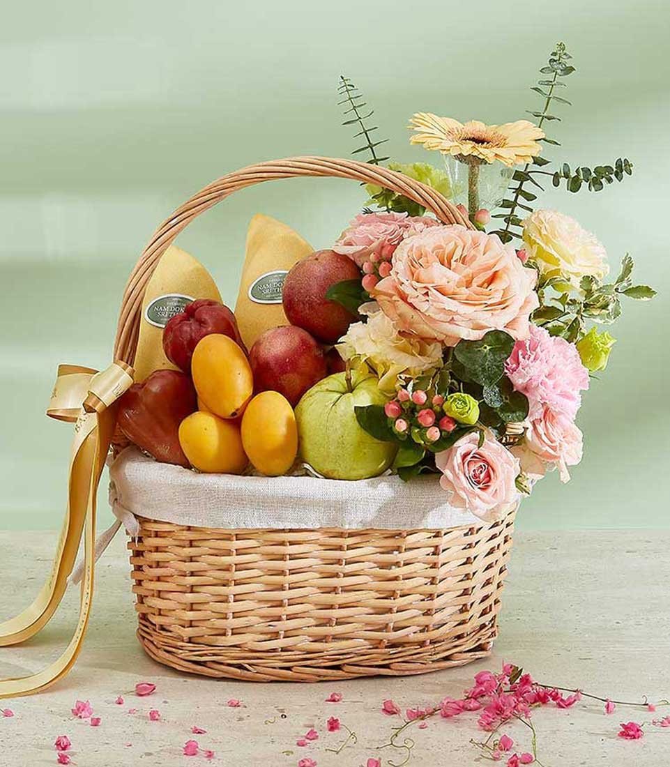 A Thai Temptations Fruit Basket is beautifully woven and filled with a variety of fresh fruits, including apples and mangoes. Adorned with pink and yellow flowers, lush greenery, and accented by a gold ribbon, the basket is set against a pastel green background scattered with delicate pink petals.