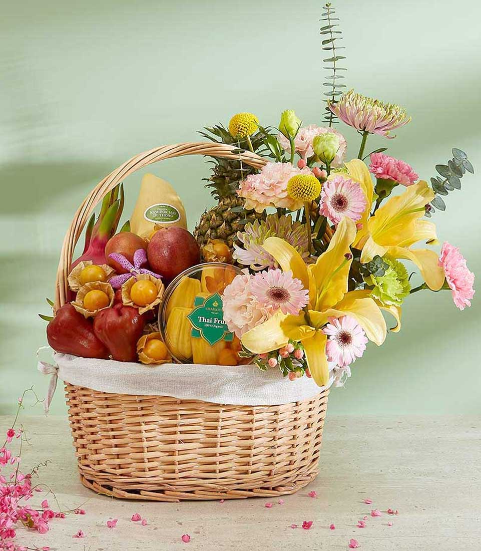 A wicker basket filled with various fresh fruits and vibrant flowers. Named the Thai Fruits Treasures Basket, it includes apples, a pineapple, and other exotic varieties. The flowers are a mix of lilies, chrysanthemums, and other blooms, creating a colorful and lively arrangement.