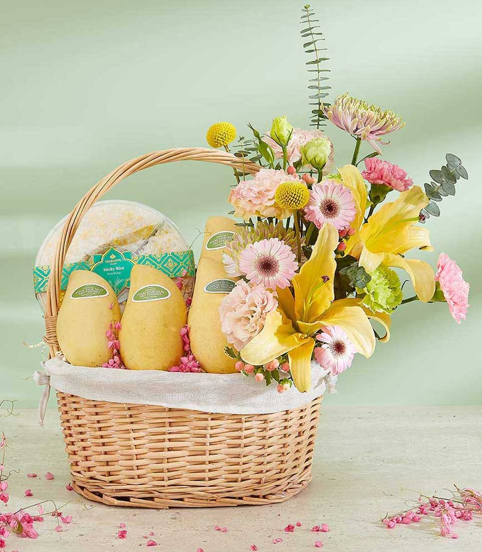 An gift basket, featuring a combination of ripe Nam Dok Mai mangoes and a package of sticky rice. The basket is adorned with a vibrant floral arrangement that includes yellow lilies, pink gerberas, and lush greenery.