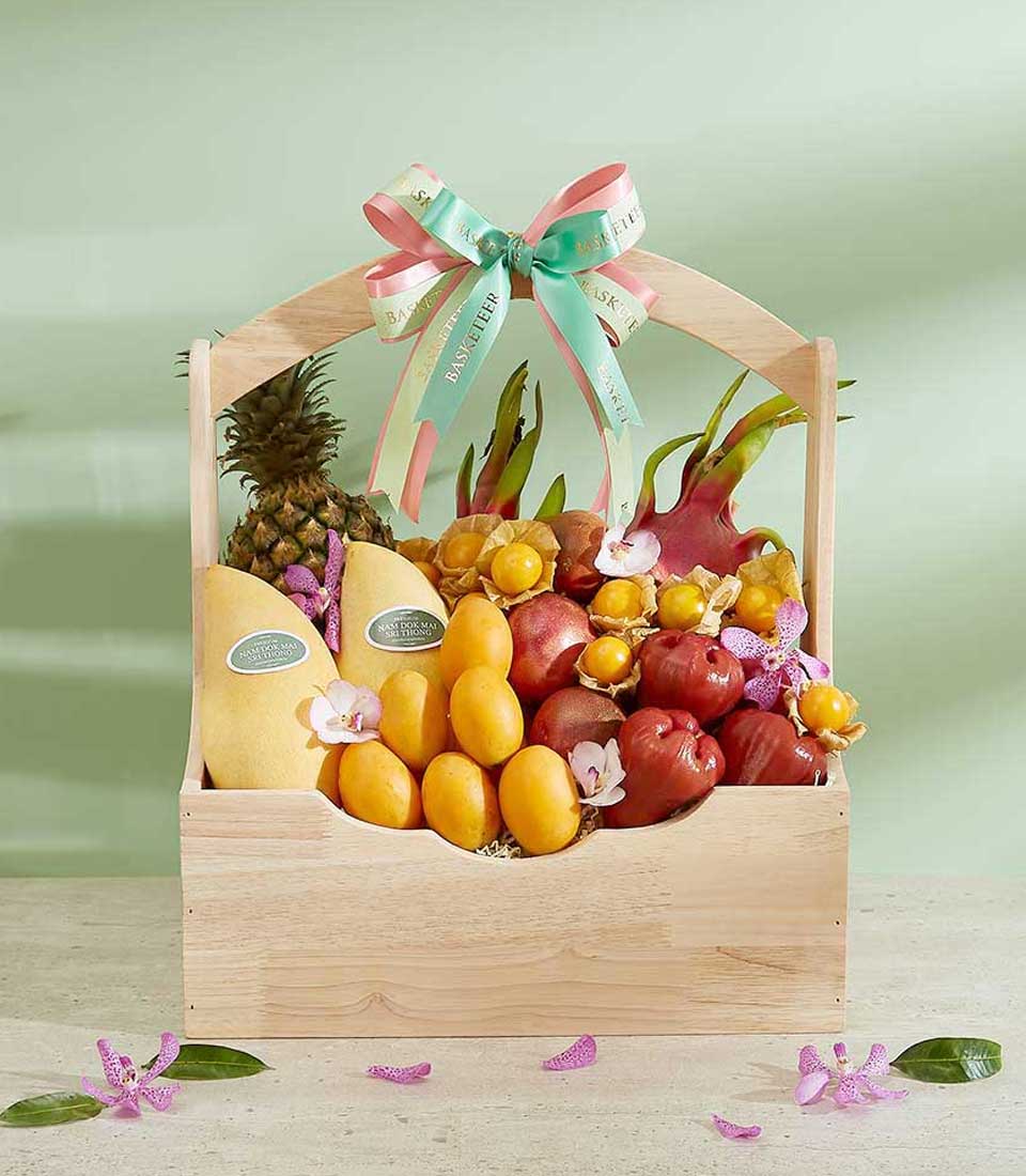 A wooden basket filled with assorted tropical fruits, including a pineapple, mangoes, dragon fruits, and apples, adorned with orchid flowers. The 