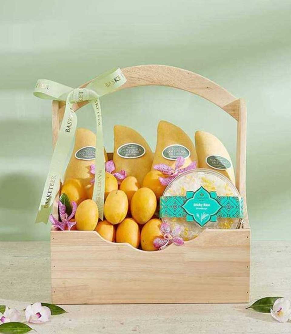 The Mango Mayongchid Delight Basket features a wooden basket filled with yellow mangoes and a green box of dried mango slices, adorned with a light green ribbon bow. The basket sits on a light-colored surface, with pink and white flowers scattered around, against a pale green background.