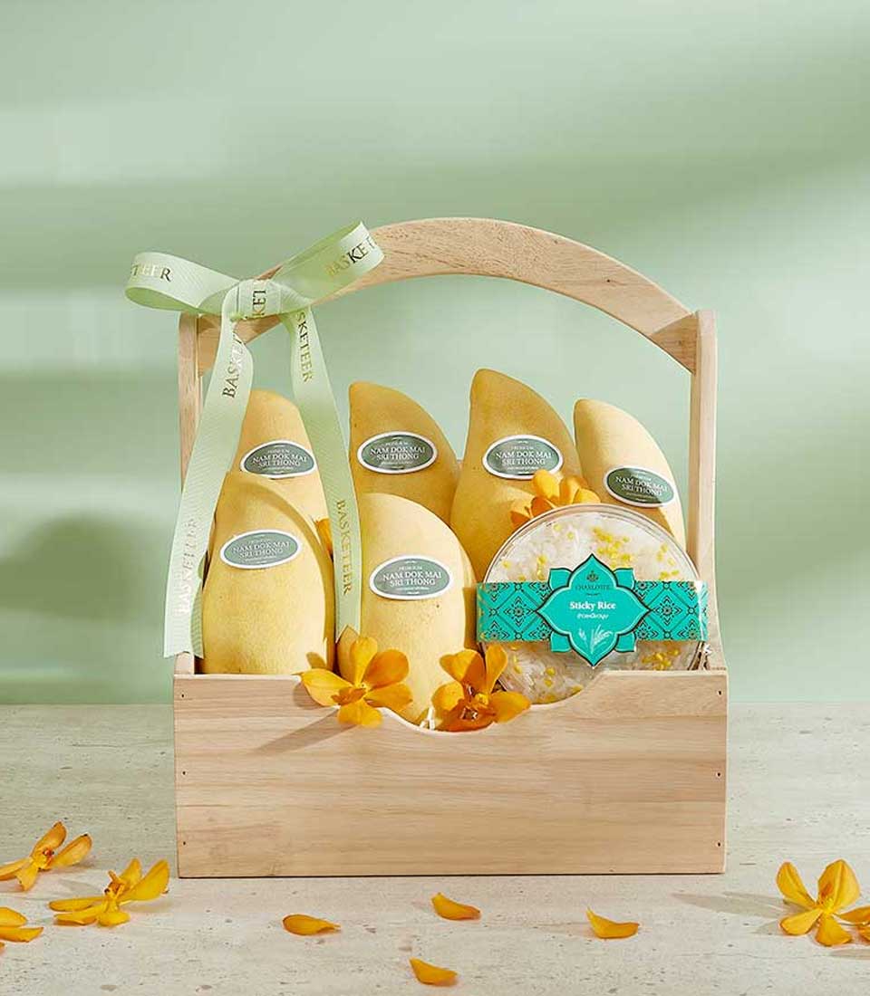 A wooden basket labeled as the Vibrant Si Thong Mango Basket contains five yellow mangoes neatly arranged with each wrapped individually and adorned with a ribbon. Beside the mangoes is an intricately designed green circular package. Yellow flower petals are scattered around the basket.