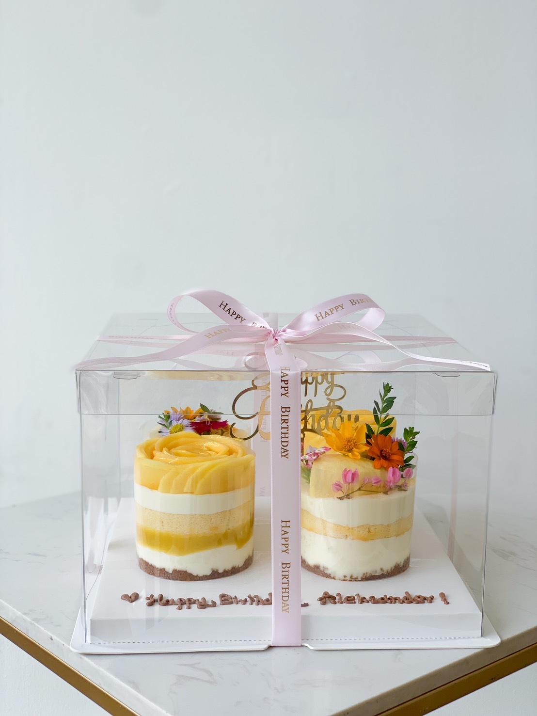 A clear box with a pink ribbon contains two Mixed Tropical Bliss Mini Layer Cakes adorned with yellow and orange flowers on top. Each cake has distinct layers of white and yellow frosting. 