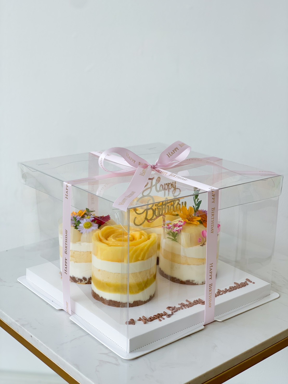 Three small Mixed Tropical Bliss Mini Layer Cakes topped with delicate yellow frosting and colorful edible flowers are displayed inside a transparent box decorated with a pink 